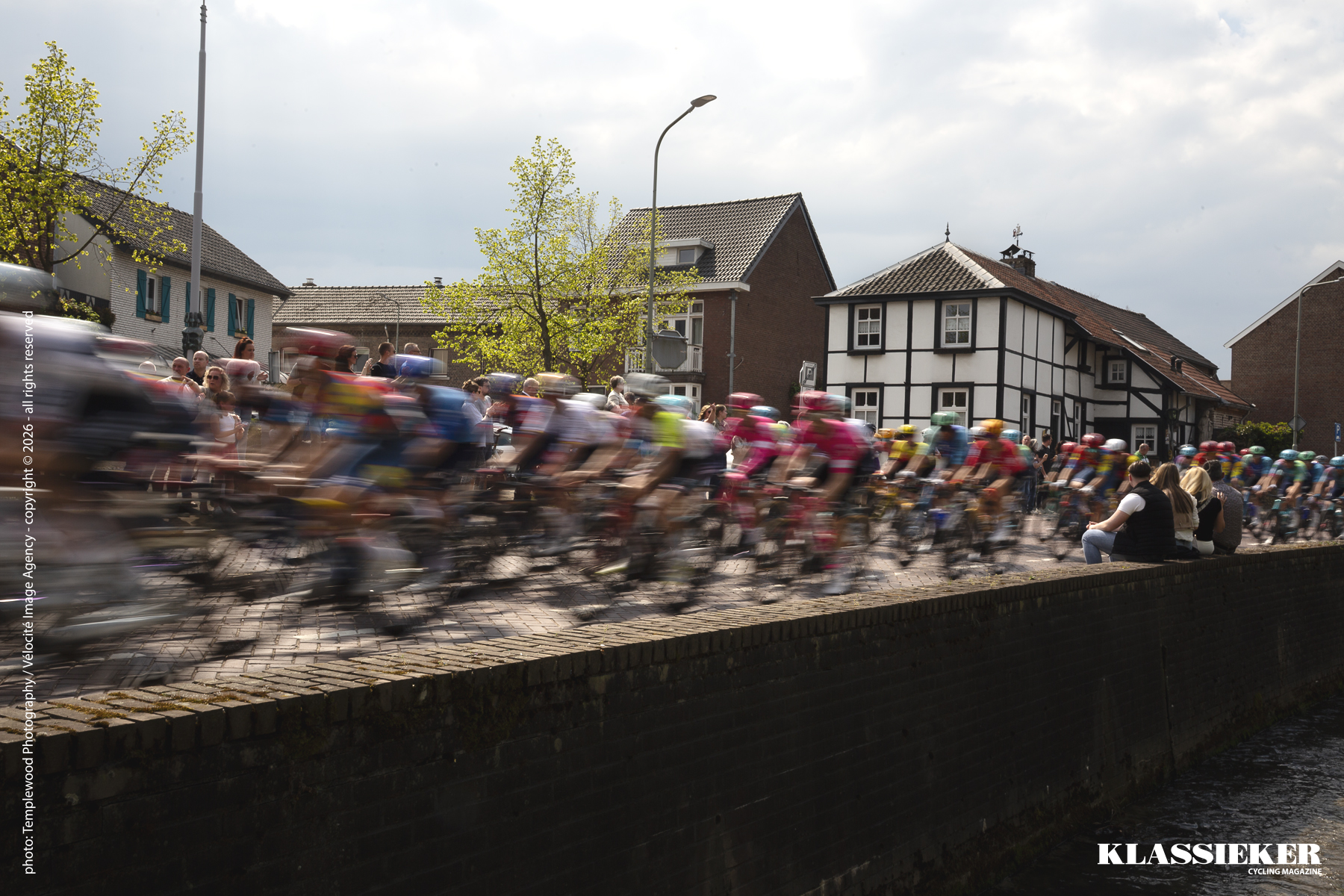Amstel Gold Race 2025 - Spectators sit on a wall close to the road as riders speed by in Mechelen