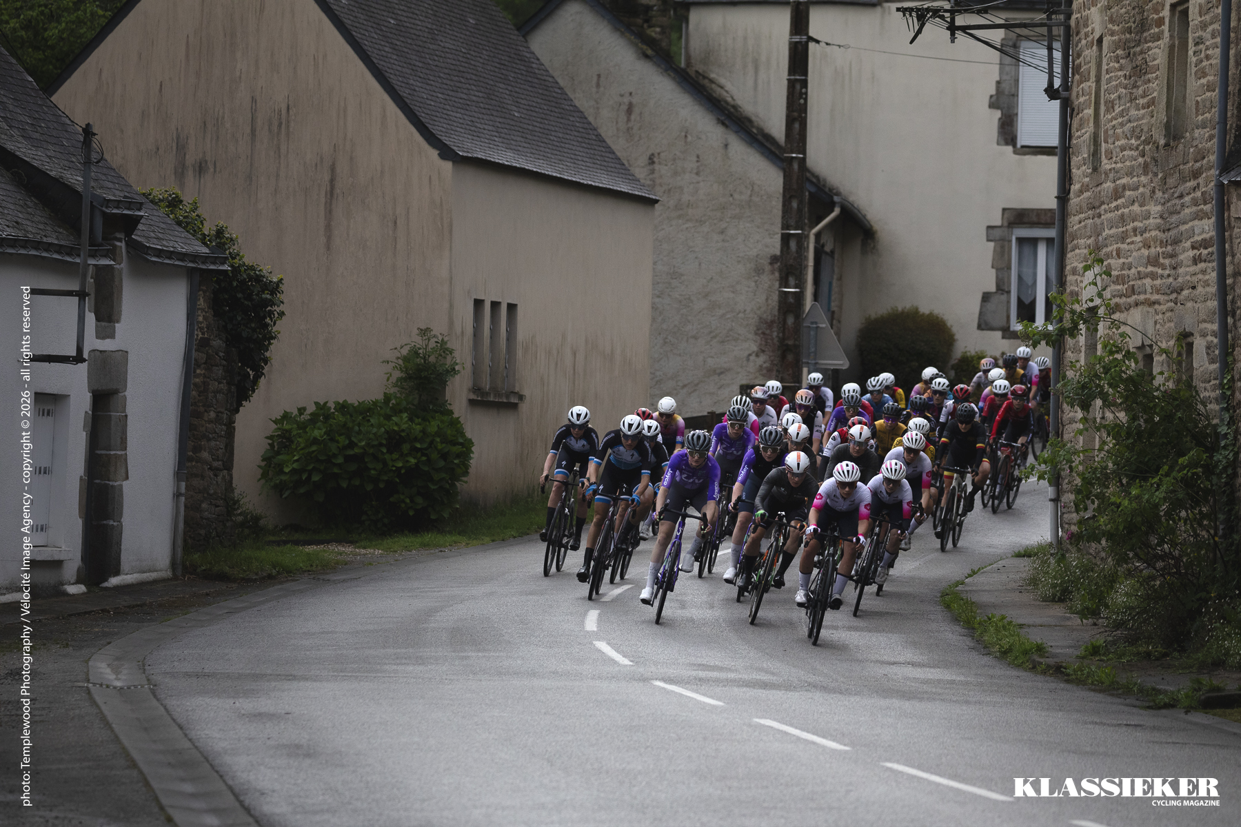 Riders round the corner, past houses at the foot of a descent in the village of Cadoudal during the 2026 Grand Prix du Morbihan Feminin 2026