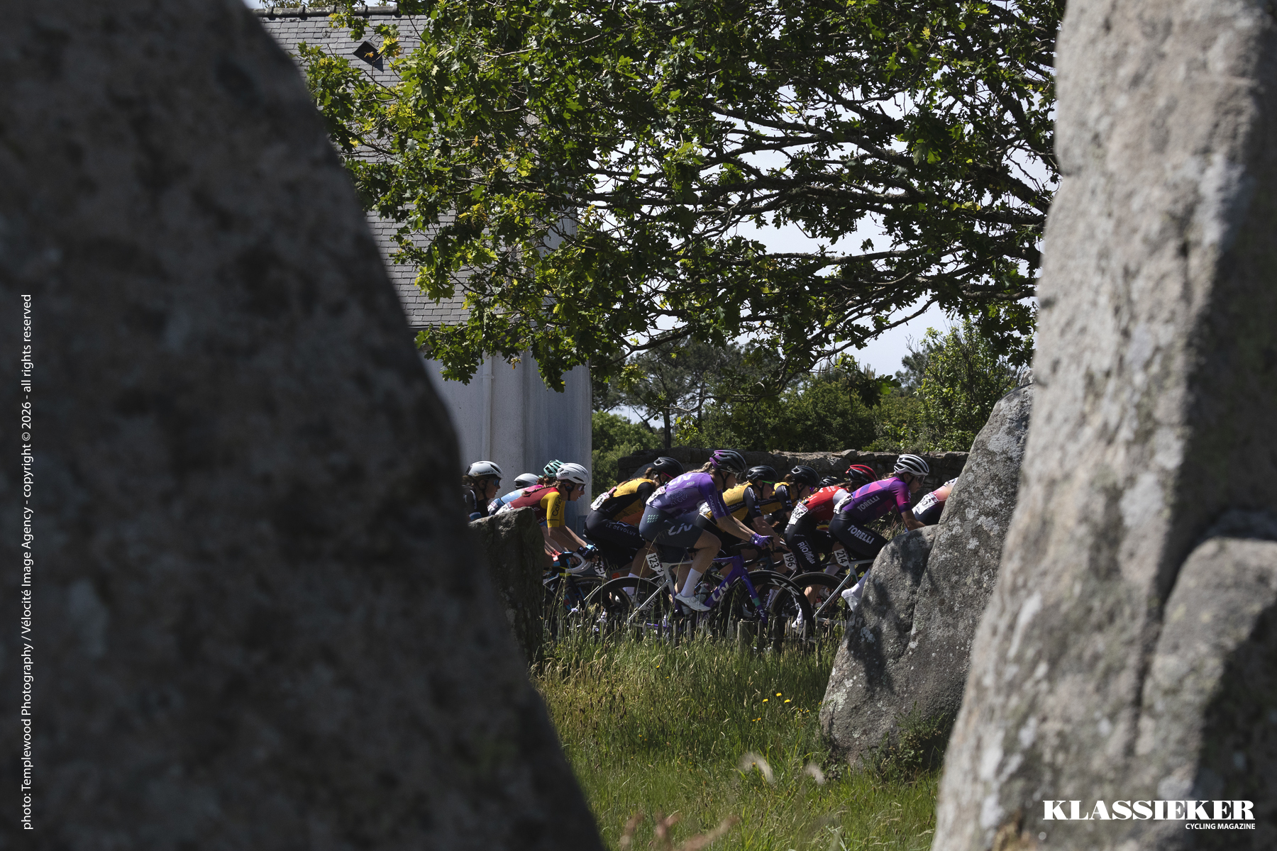 The peloton is viewed through a gap in one of the ancient monuments it passes through, Alignements de Kerzerho during La Classique Morbihan 2026