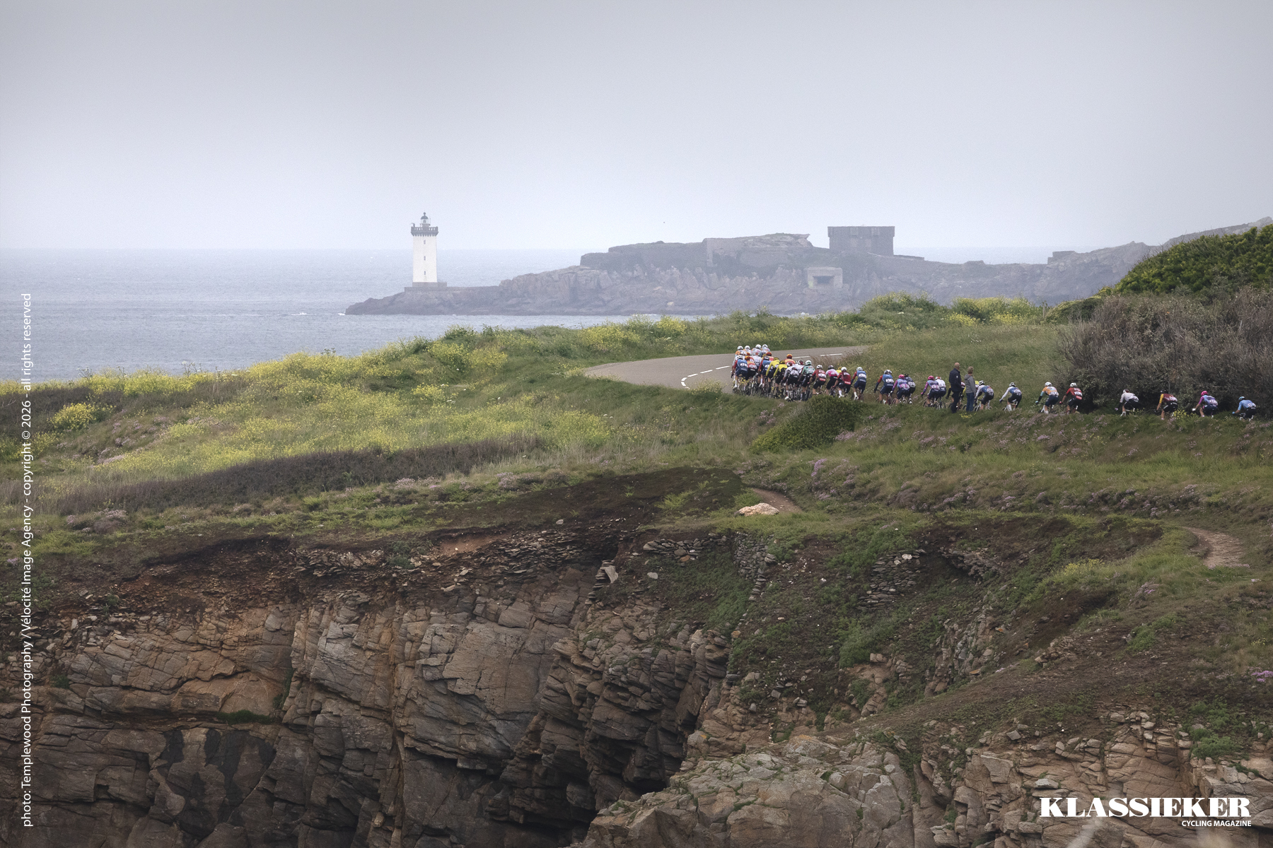 Tro Bro Leon 2026 - The peloton rounds a corner in the landscape. Cliff faces in the foreground and a white lighthouse across the sea in the distance