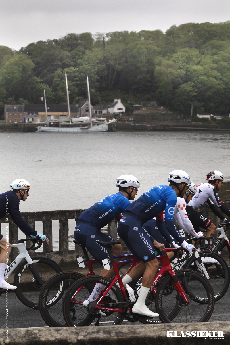 Tro Bro Leon 2026 - Riders with the sea and a sailing boat moored in the water behind them